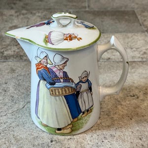 May include: White ceramic pitcher with a lid, featuring a handle and spout. The pitcher is decorated with a colorful illustration of three figures in traditional Dutch attire. The lid has a green trim and a small handle. A charming collectible.