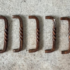 May include: Five rustic, L-shaped metal handles with a twisted, decorative center. The handles are a weathered brown color, suggesting an aged or antique appearance. They are arranged in a row against a textured, gray background.