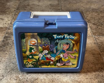 May include: Blue plastic lunchbox with a Tiny Toon Adventures theme. The lunchbox features a cartoon image of Bugs Bunny, Daffy Duck, and other characters from the show. The lunchbox is made by Thermos.