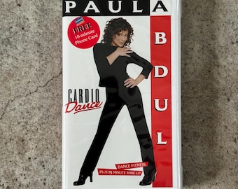 May include: A white VHS tape case featuring Paula Abdul. The case has the text "PAULA" at the top, with "CARDIO Dance" below. A photo of Paula Abdul in a black outfit is in the center. The case also says "DANCE FITNESS PLUS 15 MINUTE TONE-UP".