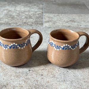 May include: Two brown ceramic mugs with handles. Each mug has a blue and white decorative pattern around the rim. The mugs are round and have a rustic appearance, suitable for coffee or tea.