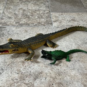 May include: A large, textured crocodile figurine with a dark green and yellow body and an open mouth showing red interior details, positioned next to a smaller, solid bright green crocodile figurine with a textured back and open mouth.
