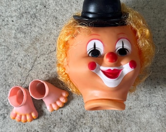 May include: A vintage clown doll head with a black bowler hat, red nose, and painted facial features. The doll has yellow hair and two detached pink feet with yellow toes. The clown's face has red cheeks and a white painted smile.