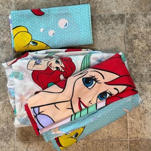 May include: Bedding set featuring a cartoon illustration of Ariel from The Little Mermaid. The set includes a light blue pillowcase with a yellow fish and white polka dots, and a white sheet with Ariel and seashells.
