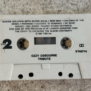 May include: A white cassette tape featuring the text "Ozzy Osbourne Tribute" and song titles such as "Iron Man" and "Paranoid." The tape is labelled "Side 2" and has a clear window to view the spools. The text "&copy; 1987 CBS Inc." is also visible.