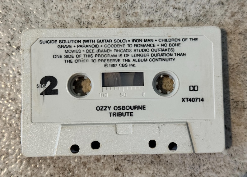 May include: A white cassette tape featuring the text "Ozzy Osbourne Tribute" and song titles such as "Iron Man" and "Paranoid." The tape is labelled "Side 2" and has a clear window to view the spools. The text "&copy; 1987 CBS Inc." is also visible.