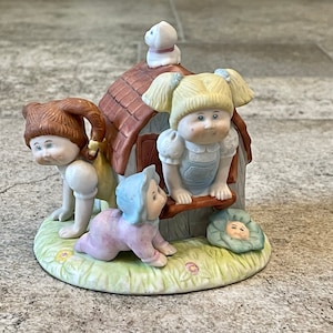 May include: A porcelain figurine set featuring three Cabbage Patch Kids dolls. The dolls are dressed in colorful outfits and are posed around a small house. The house has a brown roof and a white door. The dolls are all smiling and looking happy.