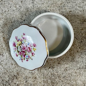 Op de afbeelding: Een witte porseleinen sieraden doos met een bloemenmotief. Het deksel heeft een gouden rand en heeft een boeket van roze, gele en witte bloemen. De doos is gesloten, maar het deksel is lichtjes open.