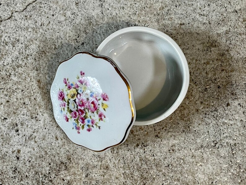 Op de afbeelding: Een witte porseleinen sieraden doos met een bloemenmotief. Het deksel heeft een gouden rand en heeft een boeket van roze, gele en witte bloemen. De doos is gesloten, maar het deksel is lichtjes open.