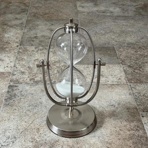 May include: A silver hourglass with clear glass bulbs and white sand. The hourglass is encased in a curved metal frame and sits on a circular base. The design is a classic timekeeping device.