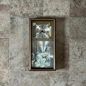 May include: Rectangular wall clock with a gold-colored frame. The top section features a clock face with black numerals and hands. The bottom section displays a painting of a deer in a forest setting.