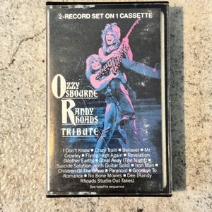 May include: A retro cassette tape for the album "Ozzy Osbourne Randy Rhoads Tribute." The cover displays a colourful image of two musicians. The tracklist includes songs such as "Crazy Train" and "Iron Man." The cassette is labelled as a 2-record set on 1 cassette.