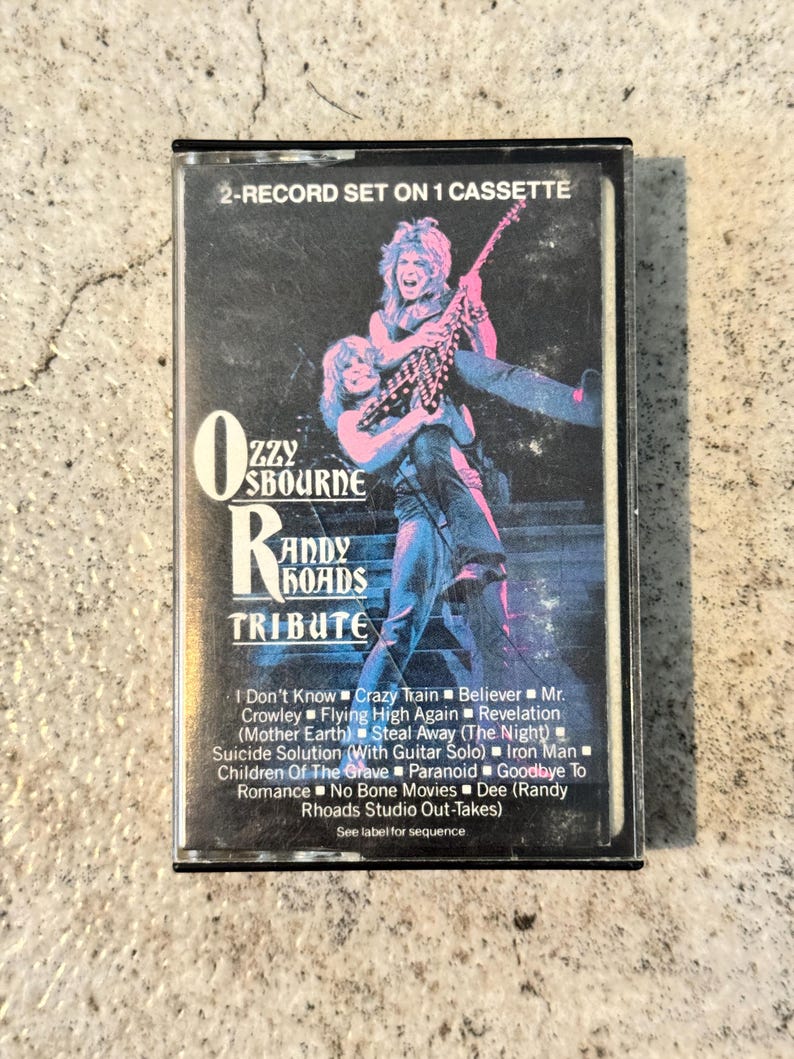 May include: A retro cassette tape for the album "Ozzy Osbourne Randy Rhoads Tribute." The cover displays a colourful image of two musicians. The tracklist includes songs such as "Crazy Train" and "Iron Man." The cassette is labelled as a 2-record set on 1 cassette.