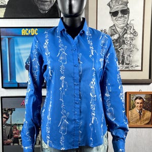 May include: A long-sleeved, button-down shirt in a vibrant blue hue, adorned with a repeating white line-art pattern of figures. The shirt is displayed on a mannequin, showcasing its design and fit. The shirt's collar is buttoned up.