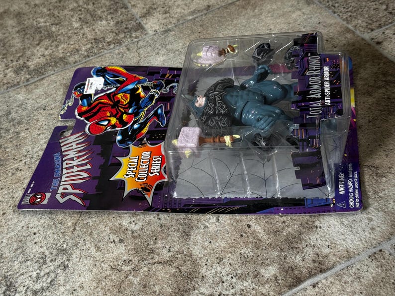 Puede incluir: Figura de acci&oacute;n de Spider-Man, "Total Armor Rhino", en un embalaje de pl&aacute;stico transparente. El embalaje es morado y negro, con las palabras "The Amazing Spider-Man" y "Special Collector Series!" impresas. La figura es azul y negra.
