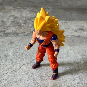 May include: A plastic action figure of a character with long, spiky yellow hair, wearing an orange martial arts uniform with a dark blue undershirt, dark blue wristbands, and dark blue boots with red accents, standing in a forward-leaning pose.
