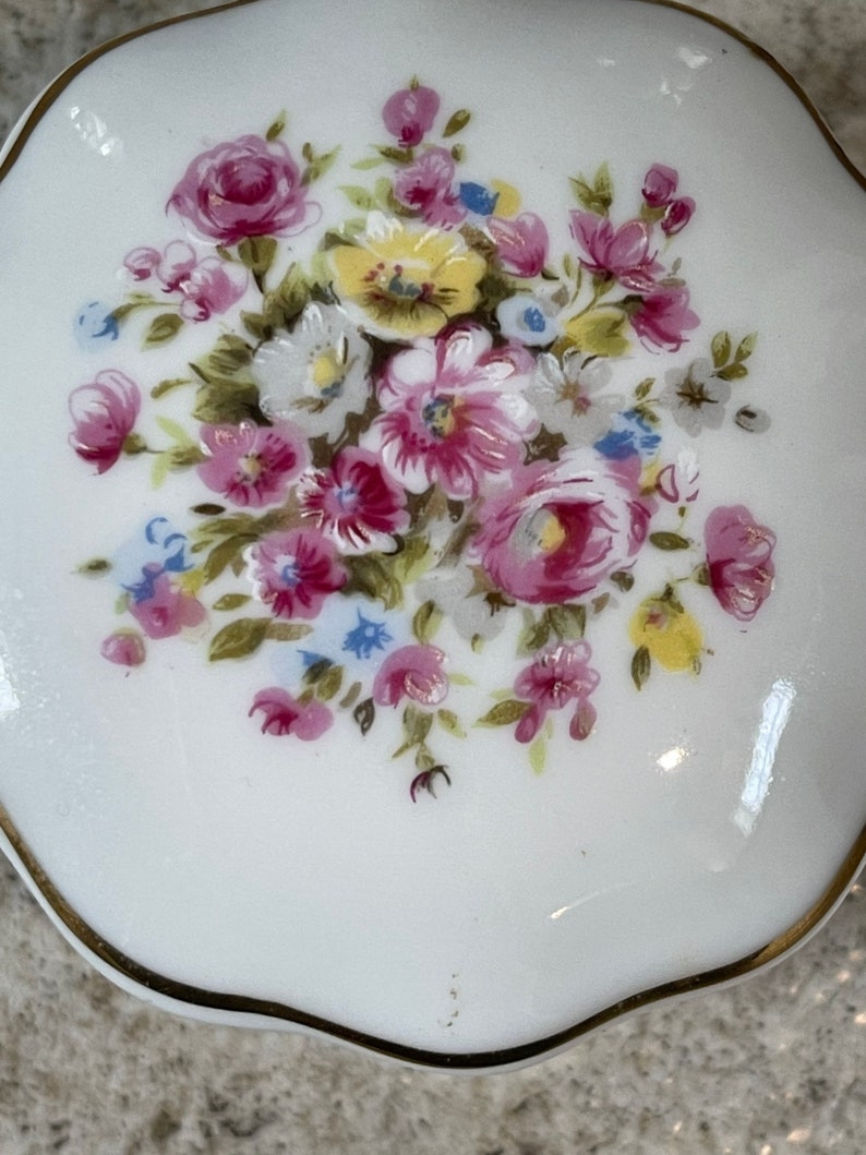Op de afbeelding: Een witte porseleinen sieraden doosje met een gouden rand en een bloemenontwerp met roze, gele en blauwe bloemen.