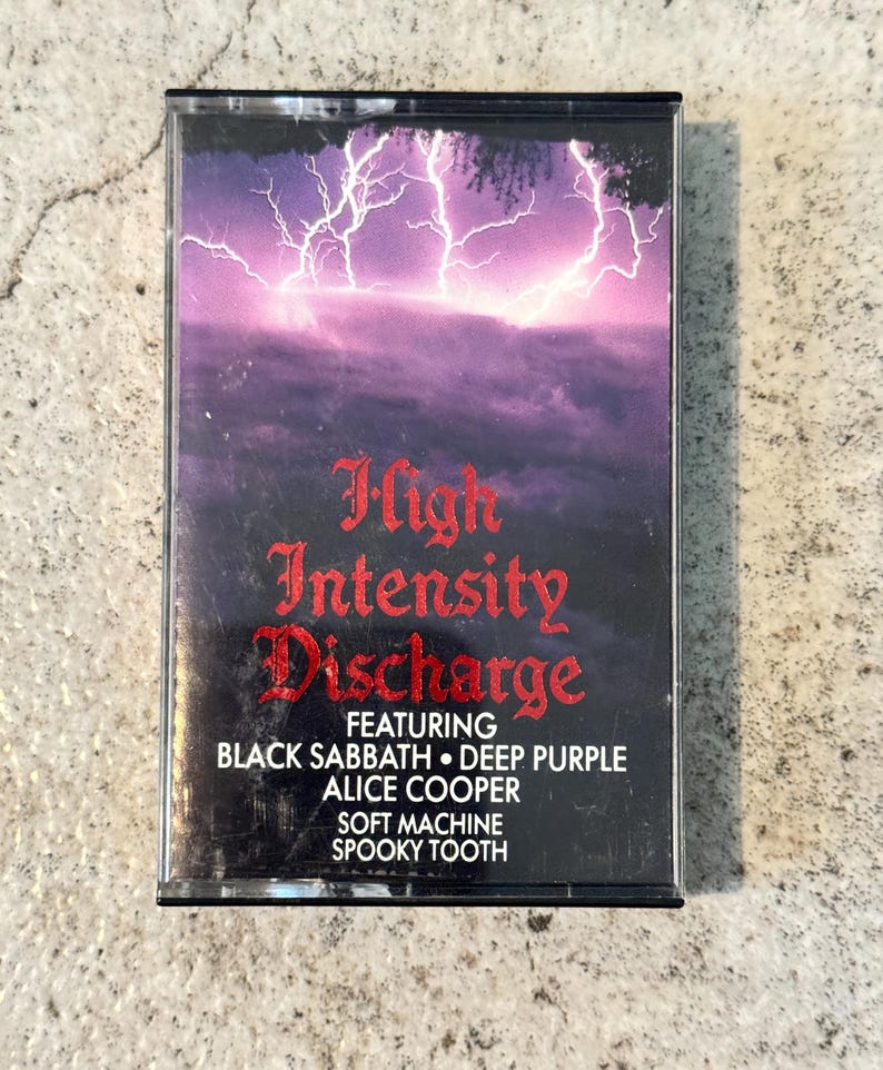 May include: A retro cassette tape with a purple and black cover featuring a lightning storm graphic. The title "High Intensity Discharge" is in red gothic lettering, with artist names like Black Sabbath and Alice Cooper.