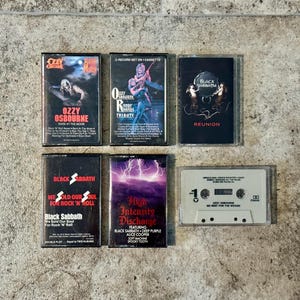 May include: A selection of vintage cassette tapes featuring rock and heavy metal bands. The tapes include albums by Ozzy Osbourne, Black Sabbath, and others. The album covers display band names, album titles, and graphic artwork. The tapes are arranged on a textured surface.