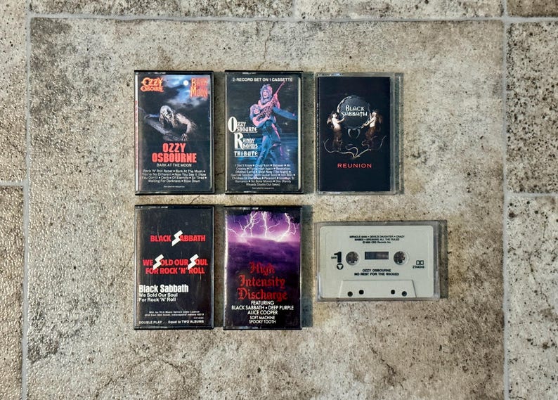 May include: A selection of vintage cassette tapes featuring rock and heavy metal bands. The tapes include albums by Ozzy Osbourne, Black Sabbath, and others. The album covers display band names, album titles, and graphic artwork. The tapes are arranged on a textured surface.
