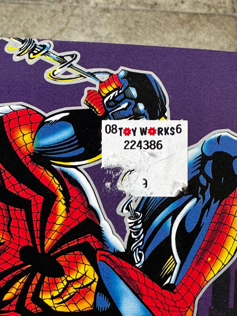 Puede incluir: Ilustraci&oacute;n de c&oacute;mic que presenta a Spider-Man en una pose din&aacute;mica. La obra de arte muestra el traje rojo y negro de Spider-Man con detalles de telara&ntilde;a, sobre un fondo p&uacute;rpura. Una pegatina con el texto "08 TOY WORKS 6 224386" es parcialmente visible.