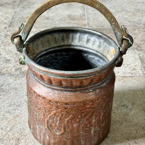 May include: A copper bucket with a brass handle. The bucket has an embossed floral pattern around the body. The handle is arched and attached to the bucket with decorative hinges. The bucket is cylindrical and has a slightly flared rim.