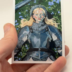 May include: A rectangular art print featuring a portrait of a person in medieval armor, with blonde hair and a serene expression. The artwork is set against a backdrop of green foliage and blue flowers, with a sword visible.