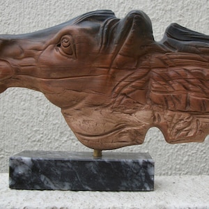 May include: A wooden sculpture of a horse's head with a textured, natural wood finish. The sculpture is mounted on a black marble base.