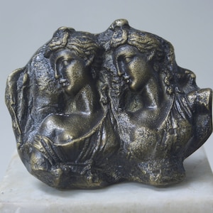 Può includere: Una scultura in bronzo di due busti di donne, con capelli e vestiti dettagliati, su uno sfondo ruvido e testurizzato.