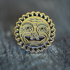 May include: Gold-colored metal pin with a black outline of a smiling sun face. The sun has a wrinkled forehead and a wide, toothy grin.