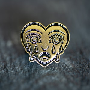 May include: Gold heart-shaped pin with a black outline and black tear-shaped drops coming from the eyes. The heart has a sad expression with a slight smile.
