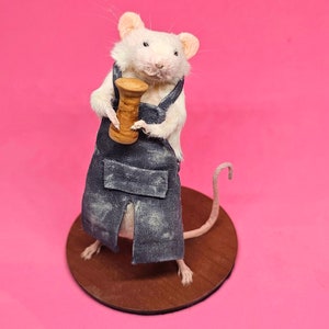 May include: A taxidermied white mouse wearing a denim apron and holding a wooden spool. The mouse stands on a round, wooden base against a bright pink background. The mouse's tail curves upwards.