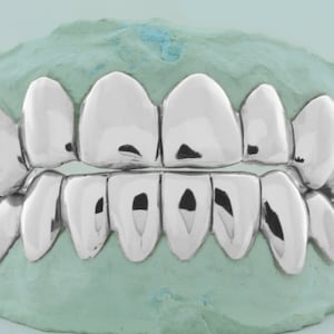 Peut inclure: Un ensemble de grilles dentaires argentées, également appelées grillz buccales, en métal. Les grillz sont conçues pour s'adapter sur les dents supérieures.