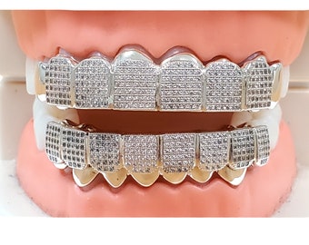 Puede incluir: Grillz plateados con una deslumbrante variedad de pequeñas piedras transparentes. Los grillz están diseñados para adaptarse a los dientes superiores e inferiores, mostrando un estilo lujoso y llamativo. Los dientes están colocados en un modelo rosa.