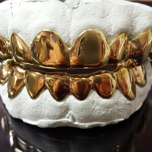 Peut inclure: Un ensemble de grillz en or, pr&eacute;sent&eacute; sur un moule dentaire blanc. Les grillz sont tr&egrave;s polis et r&eacute;fl&eacute;chissent la lumi&egrave;re, mettant en valeur leur couleur dor&eacute;e. Les dents sont dispos&eacute;es en un ensemble complet, &agrave; la fois sup&eacute;rieur et inf&eacute;rieur.