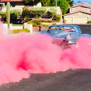 Op de afbeelding: Een zilveren klassieke auto met een roze rookwolk erachter. De auto rijdt over een woonstraat.