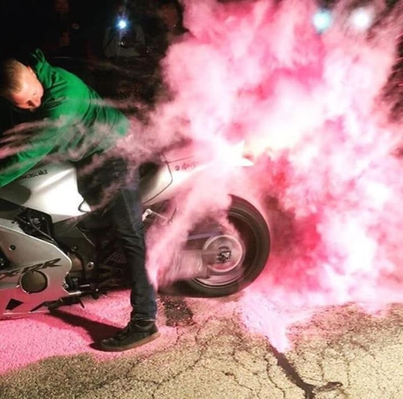 May include: A person riding a silver motorcycle with a pink smoke cloud behind it.