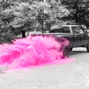 Op de afbeelding: Een zwarte pick-up truck rijdt over een geasfalteerde weg met een roze rookwolk die erachter hangt. De rookwolk is afkomstig van een gender reveal party.