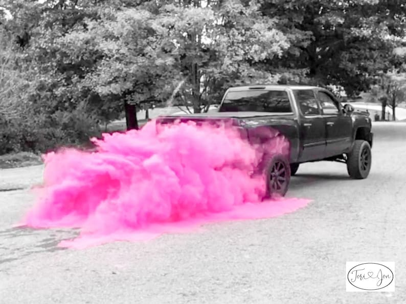 Op de afbeelding: Een zwarte pick-up truck rijdt over een geasfalteerde weg met een roze rookwolk die erachter hangt. De rookwolk is afkomstig van een gender reveal party.