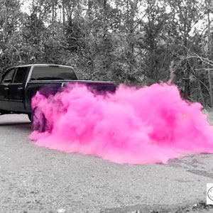Op de afbeelding: Een zwarte pick-up truck met een roze rookwolk die uit de achterkant komt. De truck rijdt op een geasfalteerde weg met bomen op de achtergrond.
