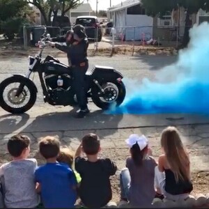 Op de afbeelding: Een zwarte motorfiets met een rijder die een zwarte helm en zwarte kleding draagt. De motorfiets laat een spoor van blauwe rook achter zich. Een groep kinderen kijkt vanaf een afstandje naar de motorfiets.
