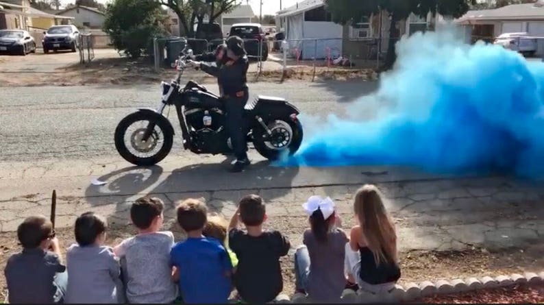 Op de afbeelding: Een zwarte motorfiets met een rijder die een zwarte helm en zwarte kleding draagt. De motorfiets laat een spoor van blauwe rook achter zich. Een groep kinderen kijkt vanaf een afstandje naar de motorfiets.