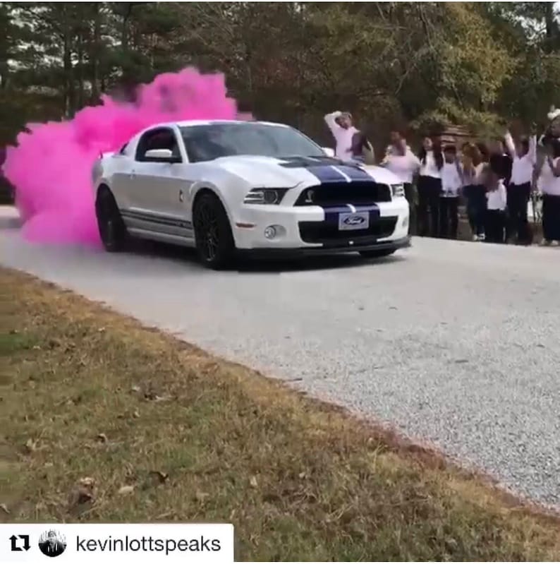 Op de afbeelding: Een witte Ford Mustang met een paarse streep rijdt over een geasfalteerde weg en laat een roze rookpluim achter zich. Een groep mensen staat aan de kant van de weg en kijkt naar de auto die voorbijrijdt.