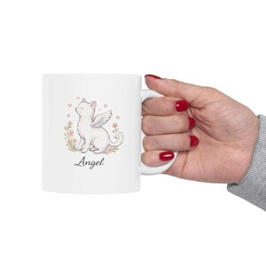 May include: White ceramic mug featuring a watercolor illustration of a white cat with angel wings, surrounded by flowers and hearts. The word "Angel" is written below the cat in a script font. The mug has a white handle.