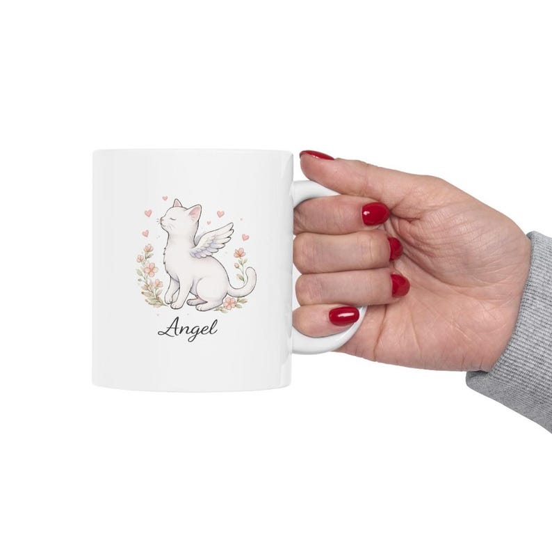 May include: White ceramic mug featuring a watercolor illustration of a white cat with angel wings, surrounded by flowers and hearts. The word "Angel" is written below the cat in a script font. The mug has a white handle.