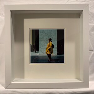 May include: A framed photograph featuring a person in a yellow coat and black backpack standing in a modern building. The image is set against a backdrop of textured walls and dark shadows, with another person in the distance.