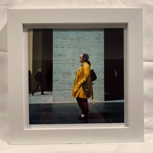 May include: A framed photograph featuring a person in a yellow coat standing in front of a textured wall. Another person is walking in the background. The photograph is in a white frame.