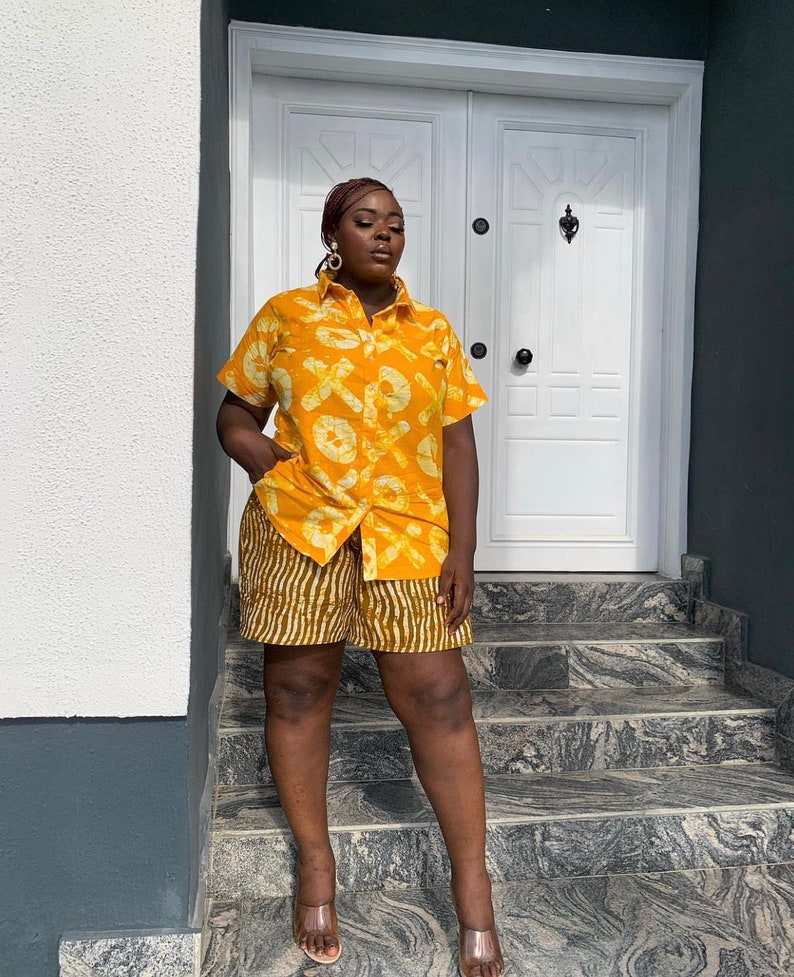 May include: A woman wearing a matching yellow button-down shirt and shorts set. The shirt and shorts feature a repeating pattern of abstract shapes. The shorts have vertical stripes. The woman is wearing clear heels and gold earrings.