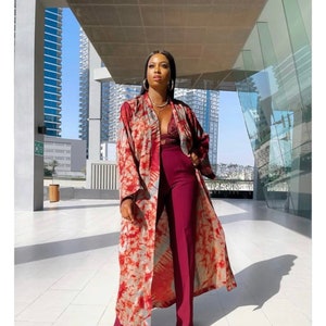 Op de afbeelding: Een vrouw draagt een lange, rood-witte tie-dye kimono met een bordeauxrode top en broek. De kimono heeft een losse pasvorm en is open aan de voorkant, waardoor de top en broek eronder zichtbaar zijn. De vrouw staat voor een modern gebouw met een glazen gevel.