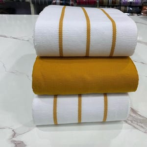 May include: Three rolls of fabric, one white with gold stripes, one solid gold, and one white with gold stripes. The fabric is folded and stacked on a white surface.